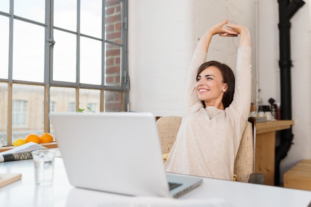 Happy relaxed young woman sitting in her kitchen with a laptop in front of her stretching her arms above her head and looking out of the window with a smile.jpeg